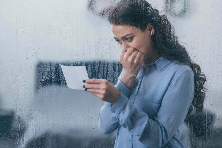 grieving woman holding photograph, covering mouth with hand and crying at home through window with